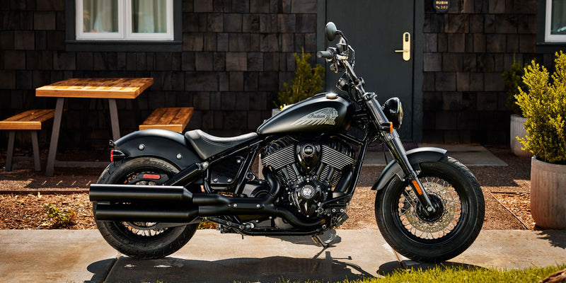 Load image into Gallery viewer, Black motorcycle parked outside a building with wooden tables and plants.