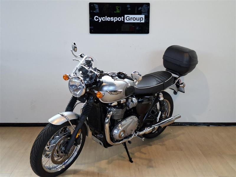 Load image into Gallery viewer, A silver 2018 Triumph Bonneville T100 motorcycle with a black seat, black hardtop storage box, and chrome details, parked indoors on a wooden floor against a white wall with a 'Cyclespot Group' sign above it.