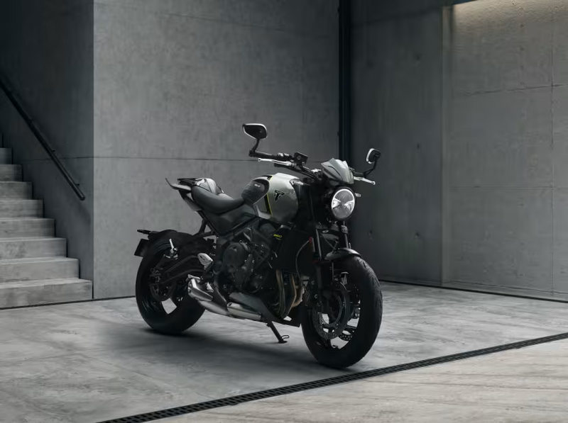 Load image into Gallery viewer, A black and silver 2026 Triumph Trident 660 LAMS motorcycle with yellow accents is parked on a concrete floor in a minimalist industrial setting with concrete walls and stairs.
