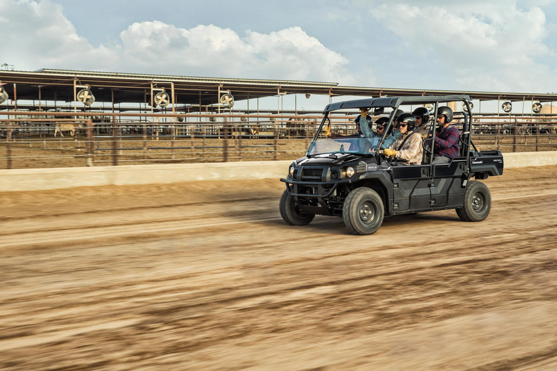 Load image into Gallery viewer, A black four-seater utility off-road vehicle with passengers driving on a dirt track, with a large covered structure in the background under a cloudy sky.