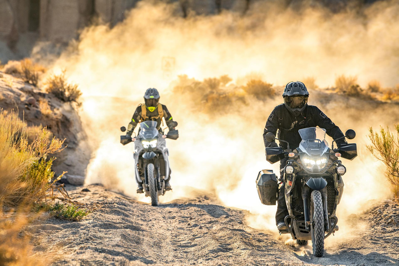 Load image into Gallery viewer, Two motorcyclists riding off-road 2026 Kawasaki KLR650 Adventure (LAMS) adventure bikes on a dusty dirt trail in a desert landscape, with one rider in the foreground and another following behind, kicking up a large cloud of golden dust.