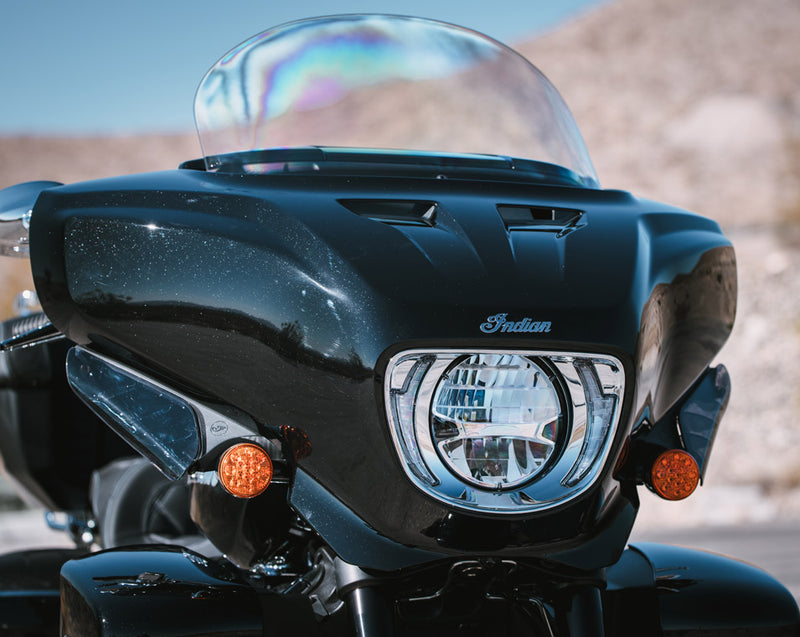 Load image into Gallery viewer, Close-up of the front of a black 2026 Indian Roadmaster Dark Horse motorcycle with a clear windshield, chrome headlight, and amber side markers, photographed outdoors against a blurred desert background.