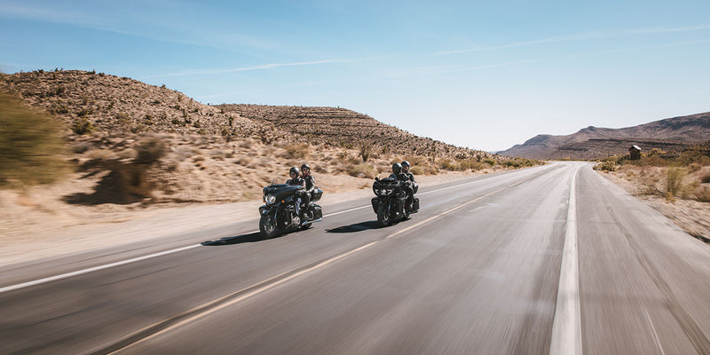 Load image into Gallery viewer, Two 2026 Indian Pursuit Dark Horse motorcycles travel side by side on a paved desert highway with rocky hills and a clear blue sky.
