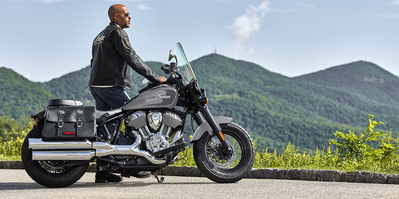 Load image into Gallery viewer, A man wearing a black leather jacket and jeans sits on a black and silver 2026 Indian Super Chief Dark Horse motorcycle with saddlebags, parked outdoors on a paved road with green mountains in the background.
