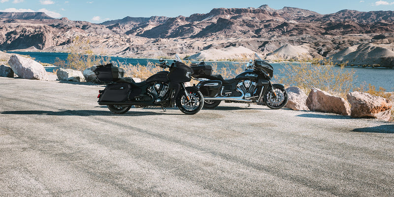 Load image into Gallery viewer, Two black 2026 Indian Pursuit Dark Horse motorcycles parked on a paved road beside a lake, with rocky mountains in the background under a blue sky.
