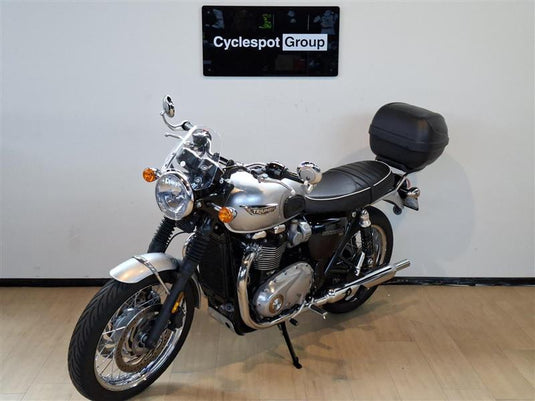 A silver 2018 Triumph Bonneville T100 motorcycle with a black seat, black hardtop storage box, and chrome details, parked indoors on a wooden floor against a white wall with a 'Cyclespot Group' sign above it.