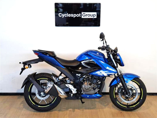 A blue and black Suzuki motorcycle displayed indoors against a white wall, with a visible 'Cyclespot Group' sign above it and a wood floor beneath.