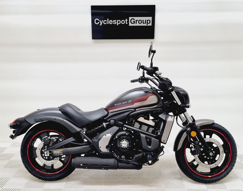 Load image into Gallery viewer, A black and dark gray Kawasaki Vulcan cruiser motorcycle with red accents on the wheels, displayed in a showroom with a 'Cyclespot Group' sign on the wall behind it.
