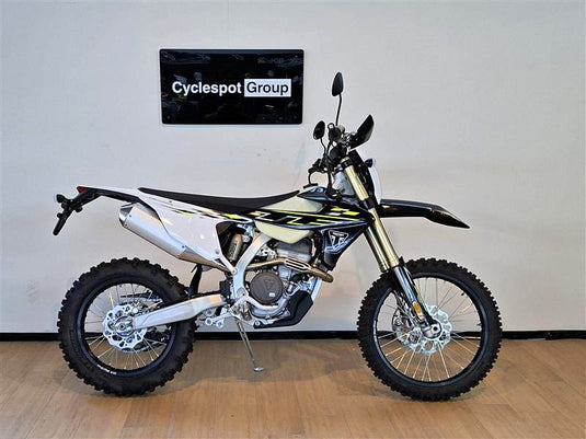 A Triumph TF 250 E dirt bike with a black, white, and yellow color scheme, displayed on a wooden floor against a white wall with a 'Cyclespot Group' sign.