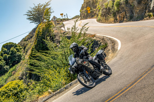 A motorcyclist wearing a black helmet and riding jacket maneuvers a 2026 Kawasaki KLR650 Adventure (LAMS) motorcycle around a sharp mountain road curve under a clear blue sky.