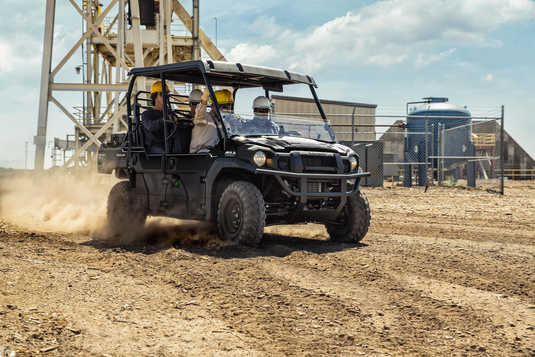 A black all-terrain utility vehicle with four passengers drives on a dusty dirt track in an industrial outdoor setting under a blue sky.
