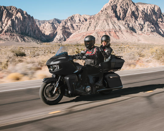 A black touring Indian Pursuit Limited with 112 Package motorcycle with two riders traveling on a desert road with rocky mountains in the background.