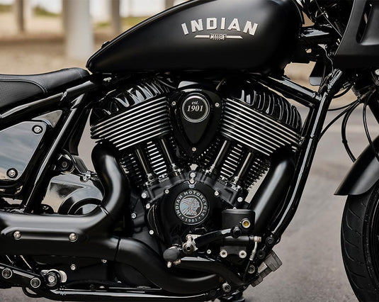 Close-up of a black Indian motorcycle engine and fuel tank with white 'INDIAN' branding on a blurred outdoor background.