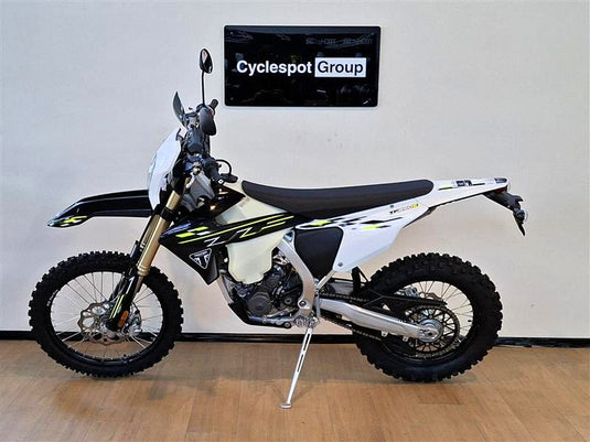 A Triumph TF 250 E dirt bike in white, black, and yellow graphics, parked on a wooden floor against a white wall with a 'Cyclespot Group' sign above it.