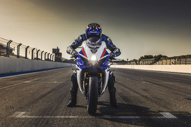 Load image into Gallery viewer, A rider in blue and white racing gear sits on a blue, white, and red Suzuki GSX-1000R 40th anniversary sport motorcycle on an open race track under a warm golden sky.
