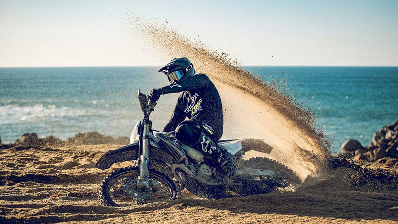 Load image into Gallery viewer, A person riding a 2026 Triumph TF 450-E dirt bike on a sandy beach with the ocean in the background, kicking up a large spray of sand.

